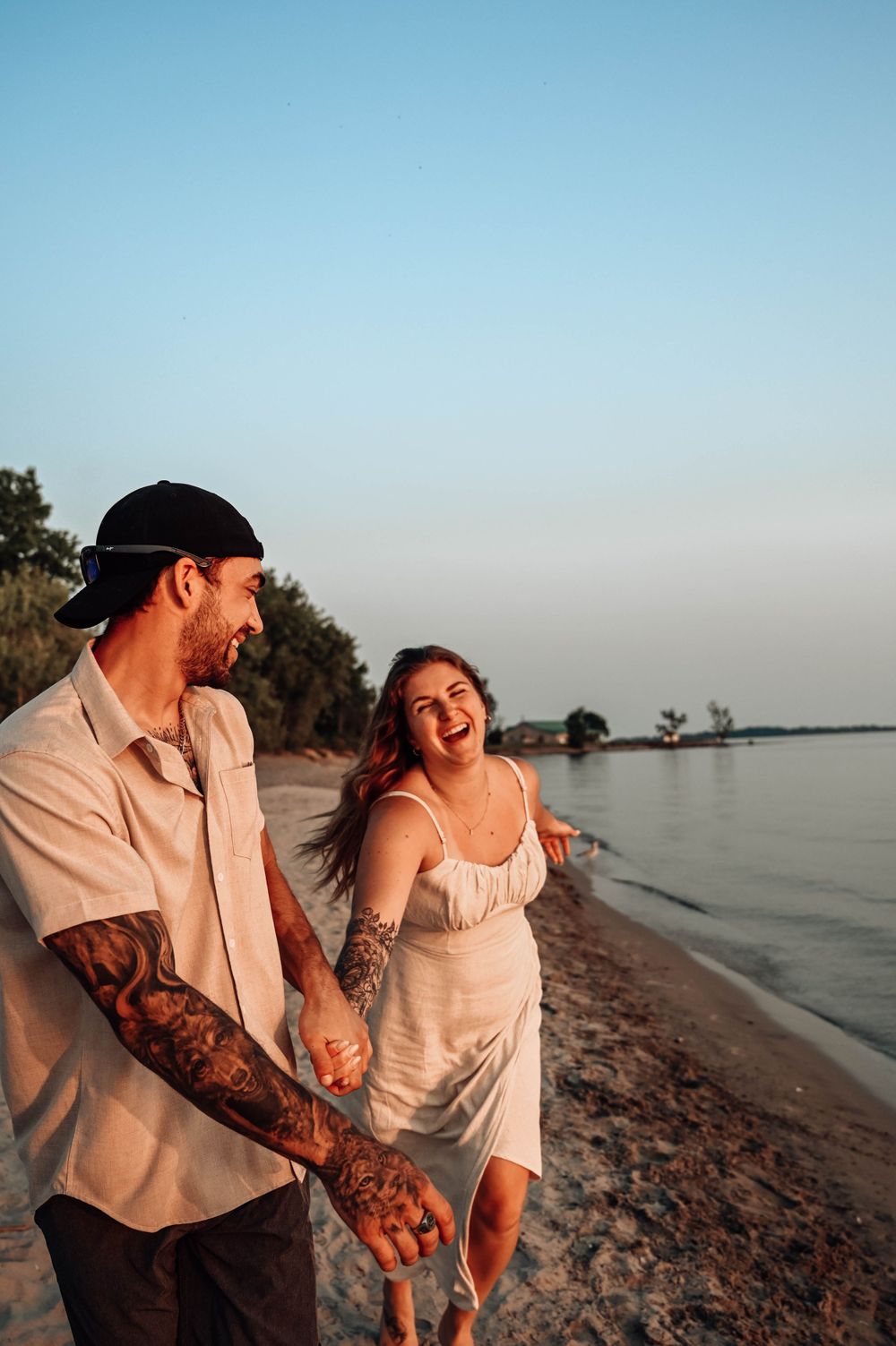Happy couple laughing and walking along the beach at sunset, with tattoos and casual summer attire.