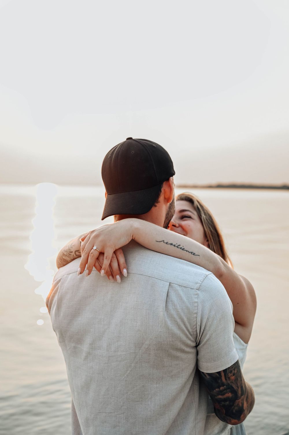 A romantic couple embraces by the water during sunset, with one person wearing a baseball cap and white shirt.