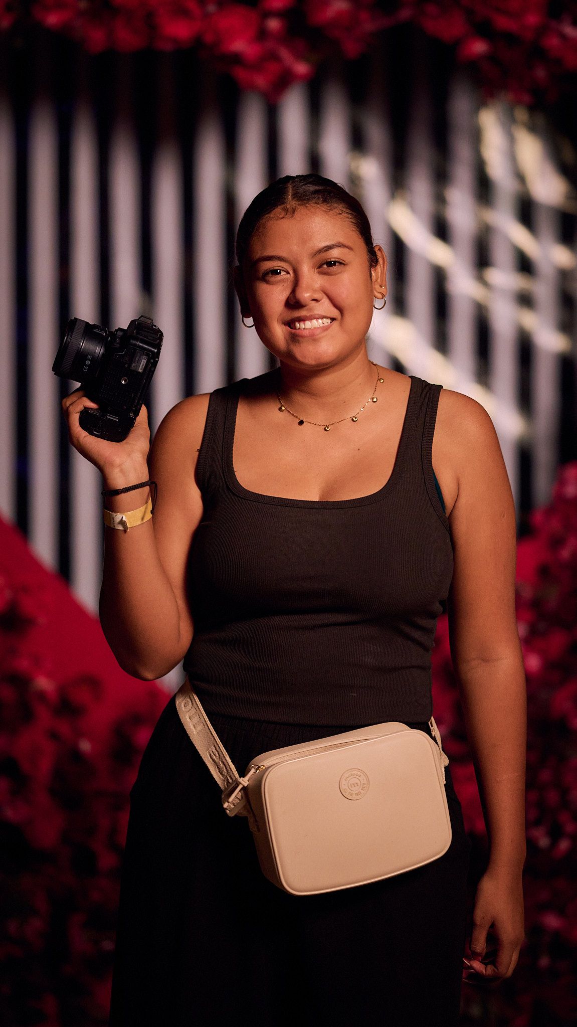 A photographer in a black tank top holds a camera while wearing a cream colored crossbody bag against a floral backdrop.