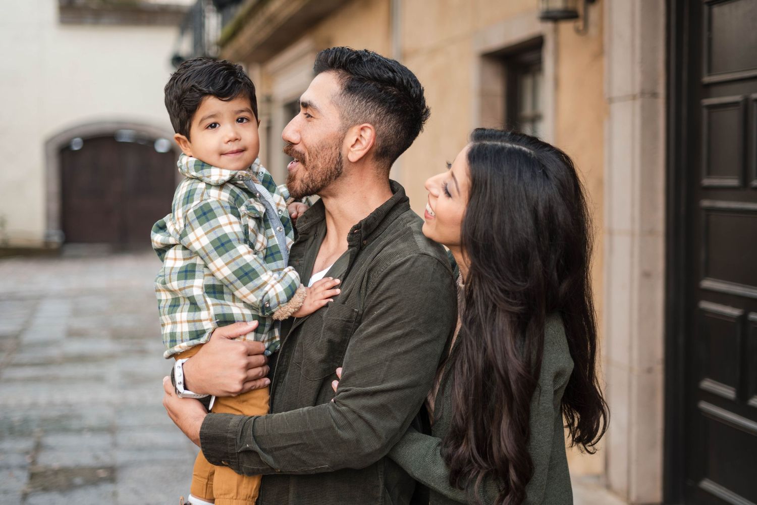 Happy Hispanic family with father holding young son while mother embraces them on a cobblestone street.