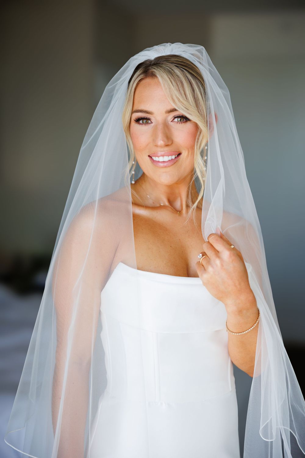 Smiling bride in white strapless gown and flowing veil holds veil edge, wearing diamond ring and earrings.