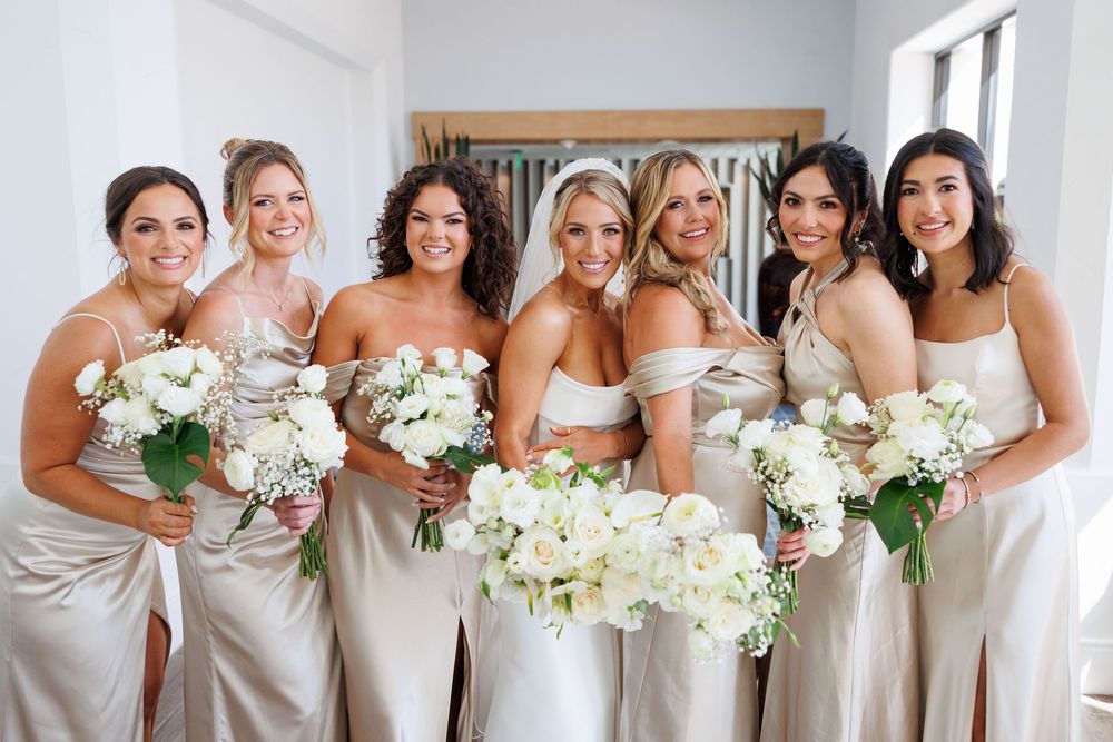 Bride and bridesmaids in champagne satin gowns holding white floral bouquets smiling together indoors.
