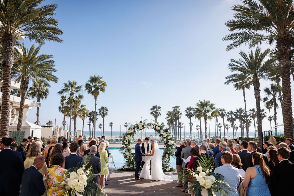 Outdoor beach wedding ceremony with couple at floral arch altar, surrounded by guests and tall palm trees under a clear blue sky.