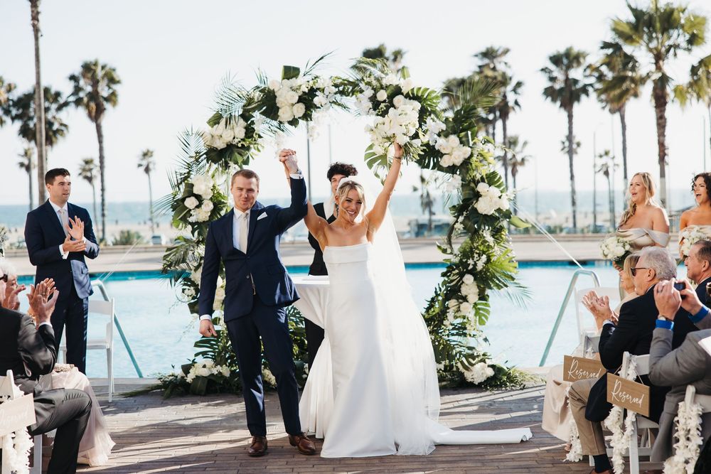 Joyful newlyweds celebrate under a floral arch at an outdoor waterfront wedding ceremony with palm trees.