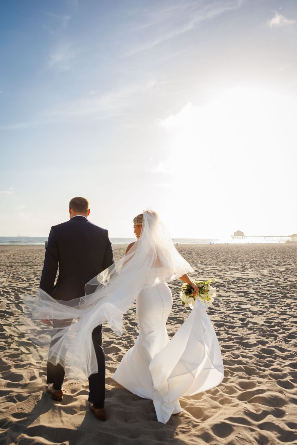 Bride and groom walking hand in hand on a sandy beach, her veil flowing in the wind, with ocean and pier in background.