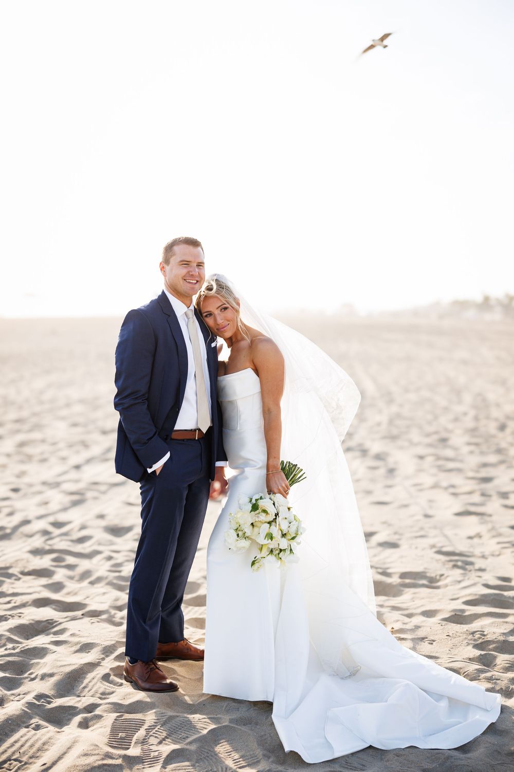 Bride and groom posing on sandy beach, bride in white gown with bouquet, groom in navy suit, sunny sky backdrop.