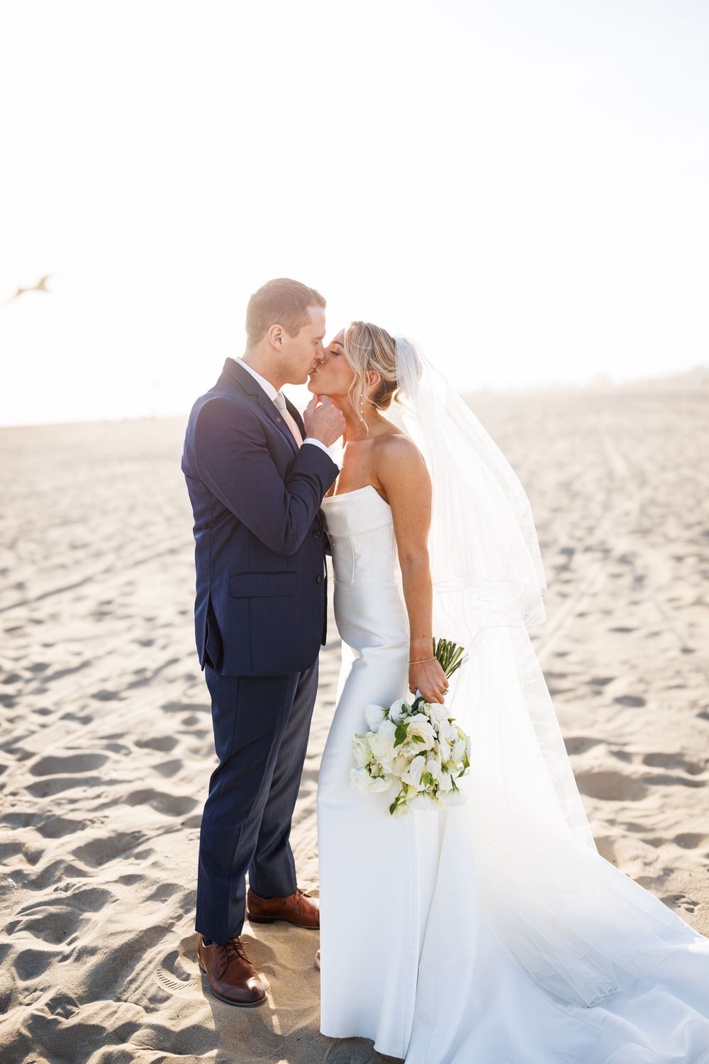 Bride and groom share a romantic kiss on a sandy beach, she holds white flowers, wearing a strapless gown and veil.