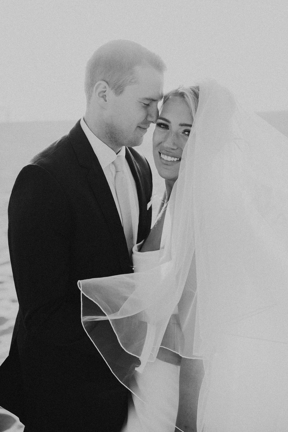 Black and white photo of a smiling bride in veil and groom in suit touching foreheads at beach wedding.