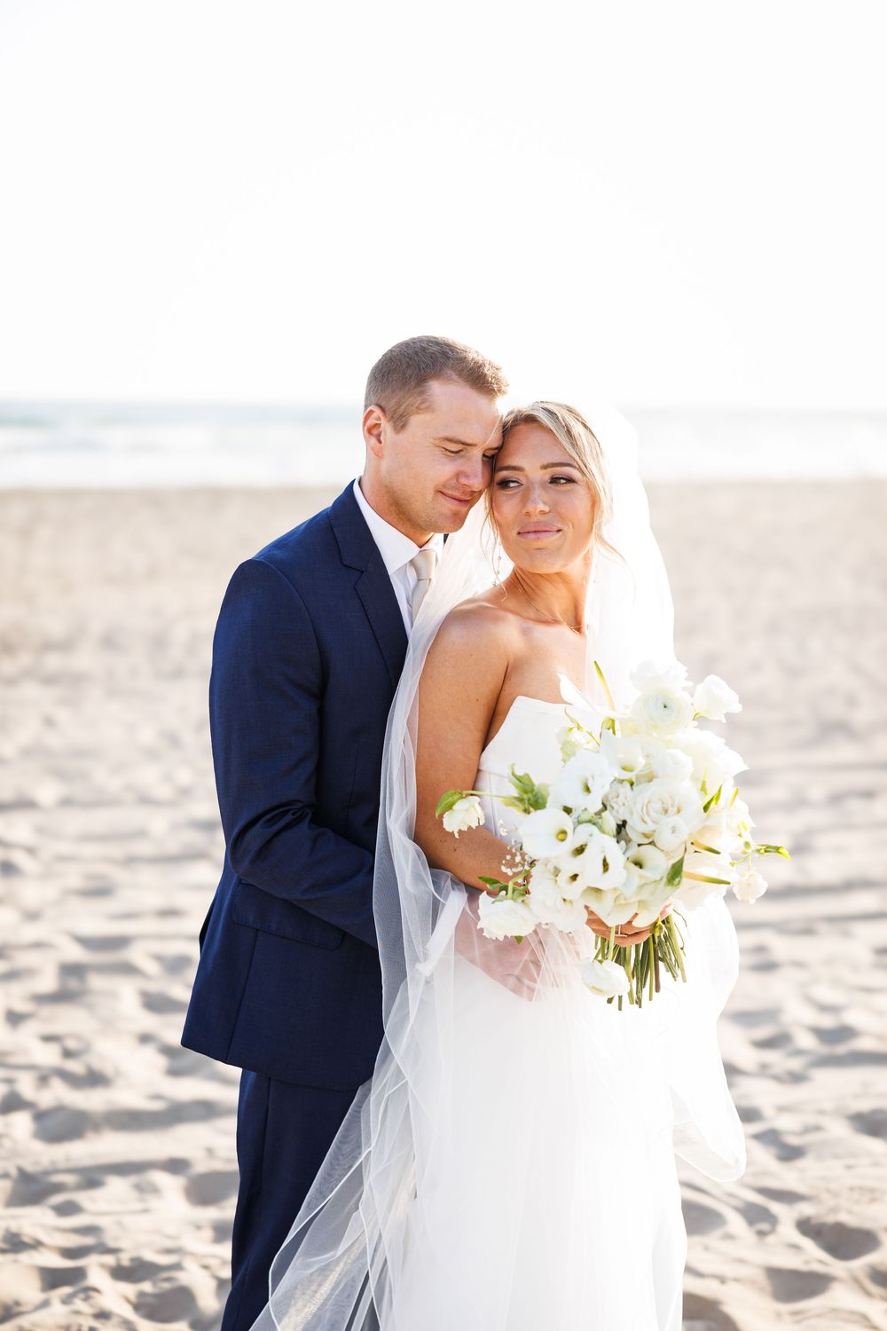 Bride holding white floral bouquet with groom embracing her on sandy beach at sunset wedding.