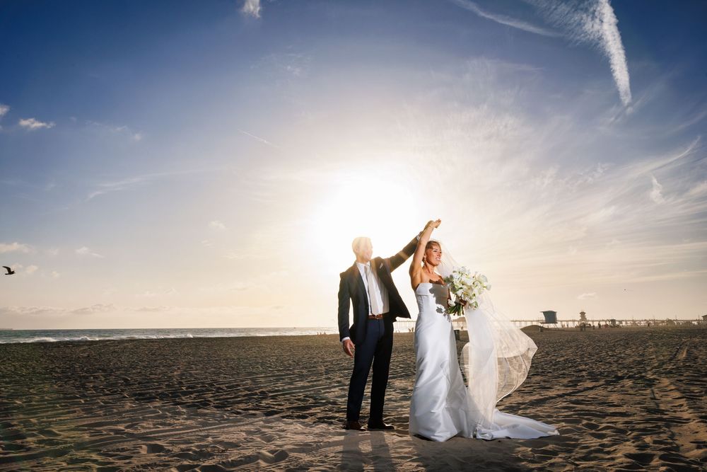 Bride and groom celebrate on a sunny beach at sunset, with veil flowing in the wind and ocean in the background.