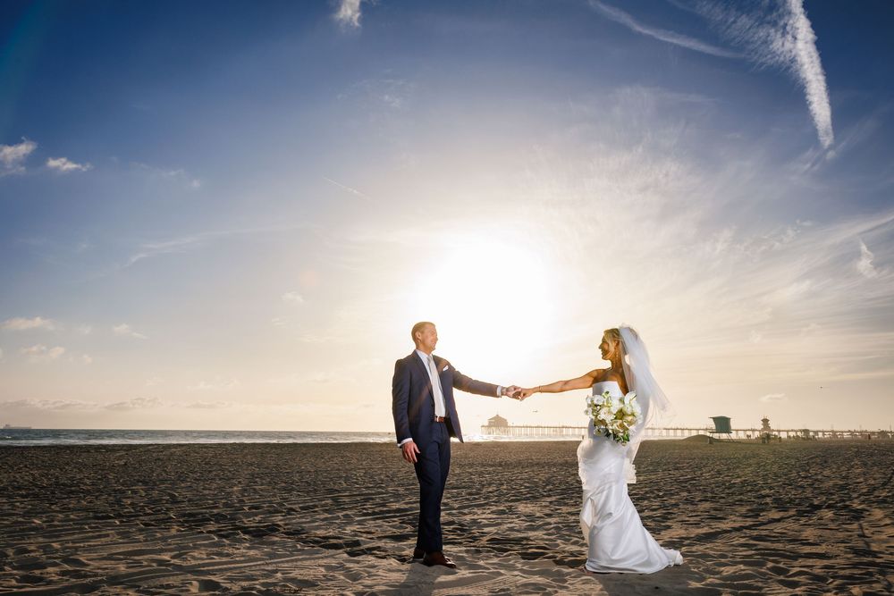 Bride and groom holding hands on sunny beach with dramatic sky at outdoor coastal wedding.