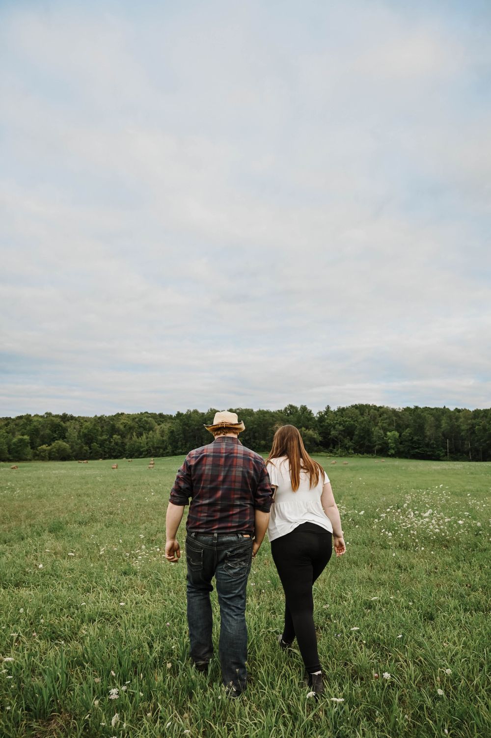 A couple walks together through a green spring meadow with trees and cloudy sky in the background.