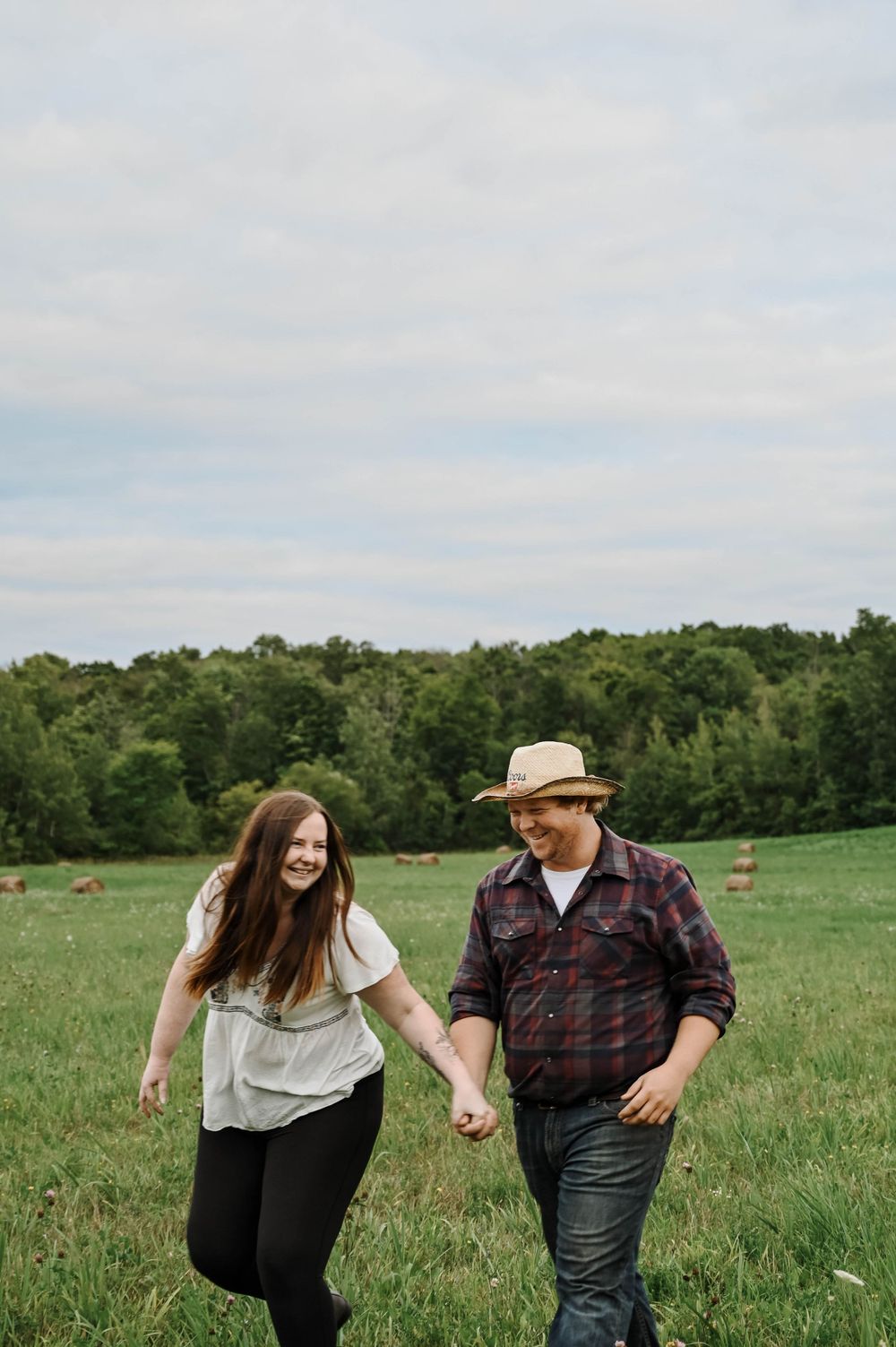 A couple holds hands while walking together through a green meadow with trees in the background on an overcast day.