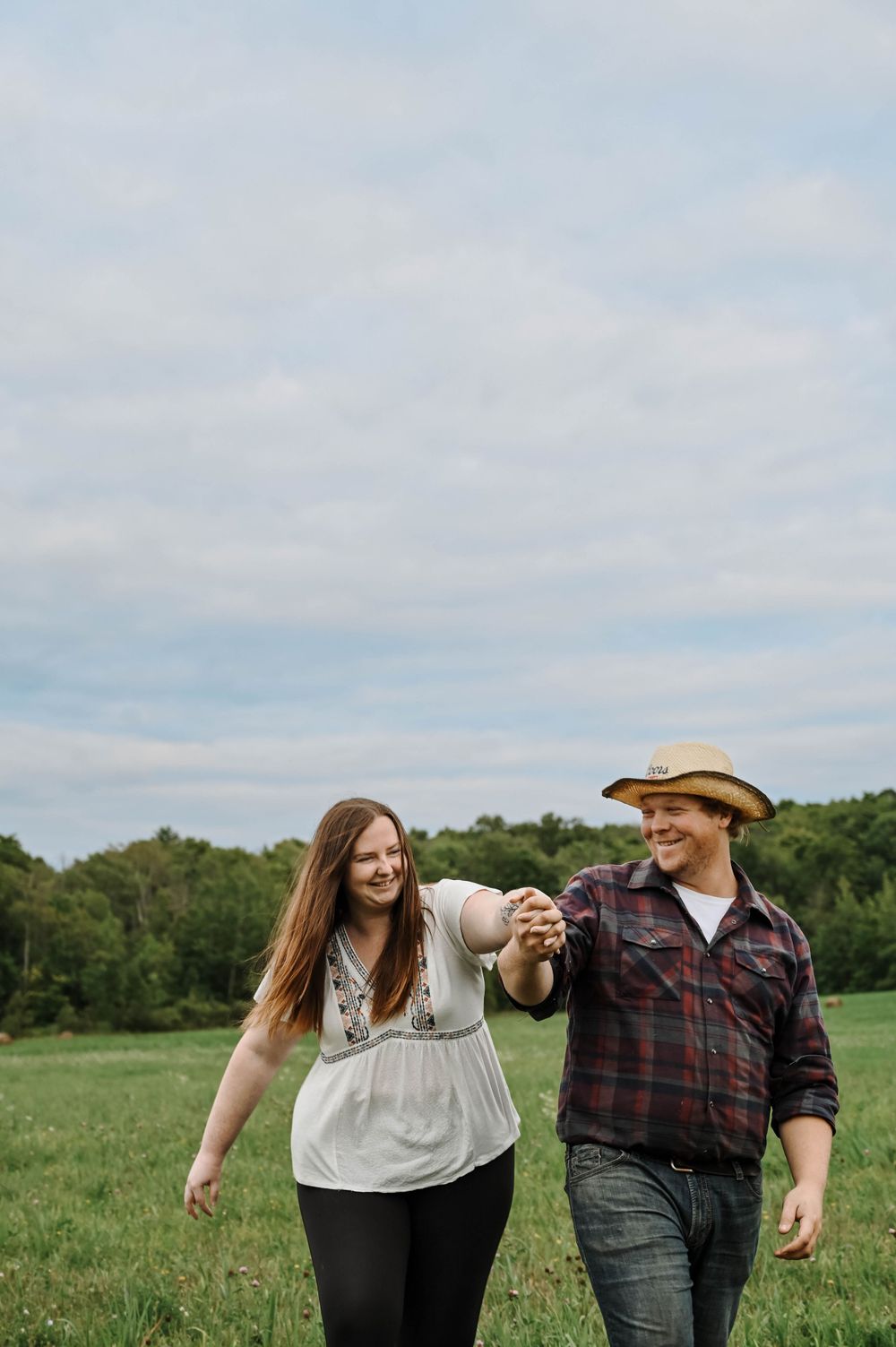 A couple holding hands while walking together through a green field on a cloudy day.
