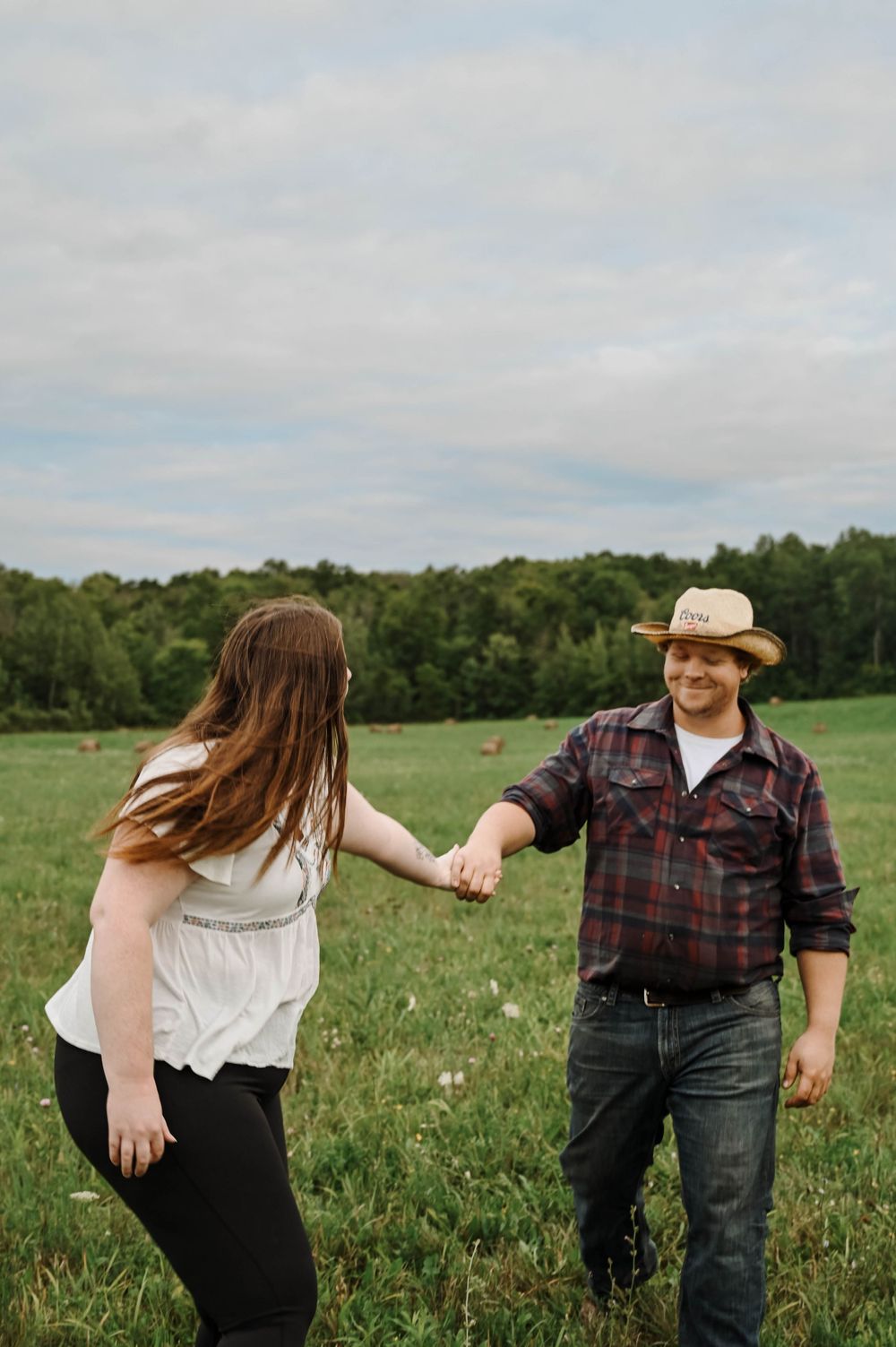 A couple holds hands while standing in a grassy meadow with trees in the background on an overcast day.