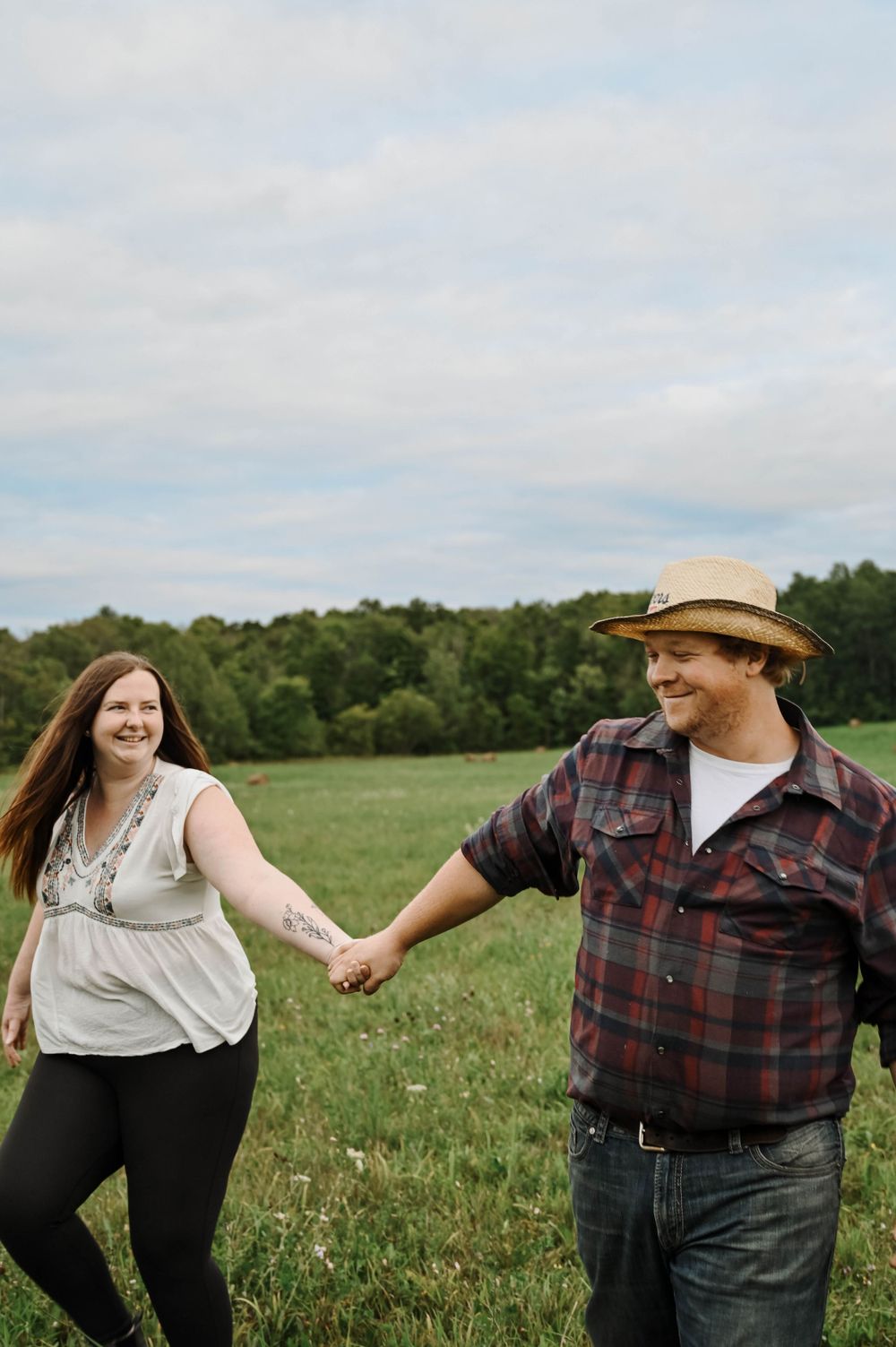 A couple holding hands and laughing together in a sunny meadow with trees in the background on a summer day.