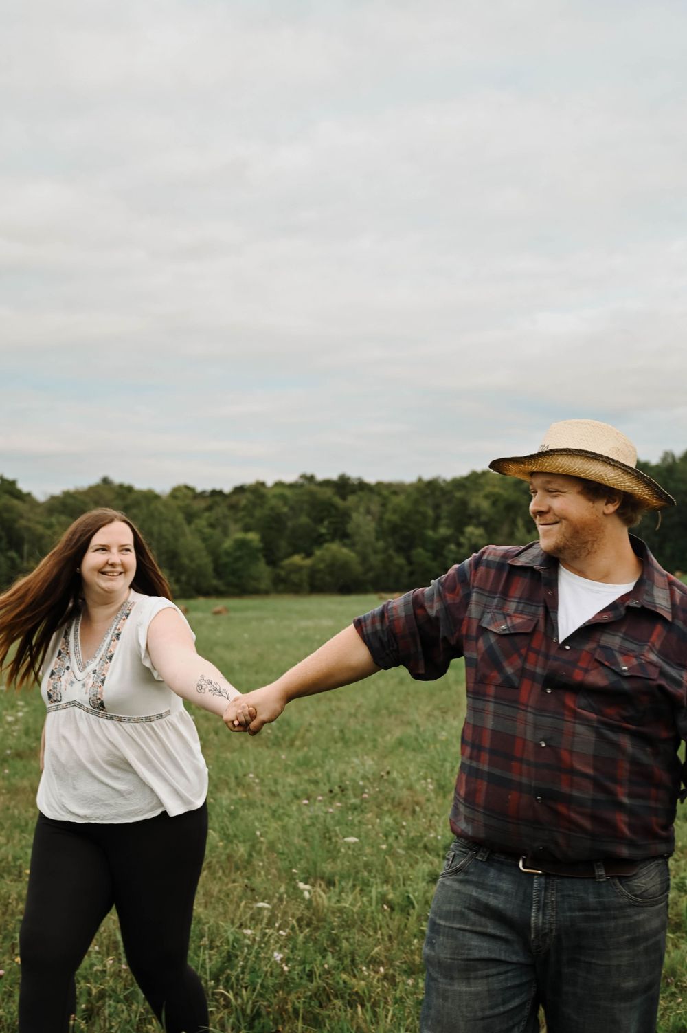 Couple holding hands and laughing in a green field on an overcast day.