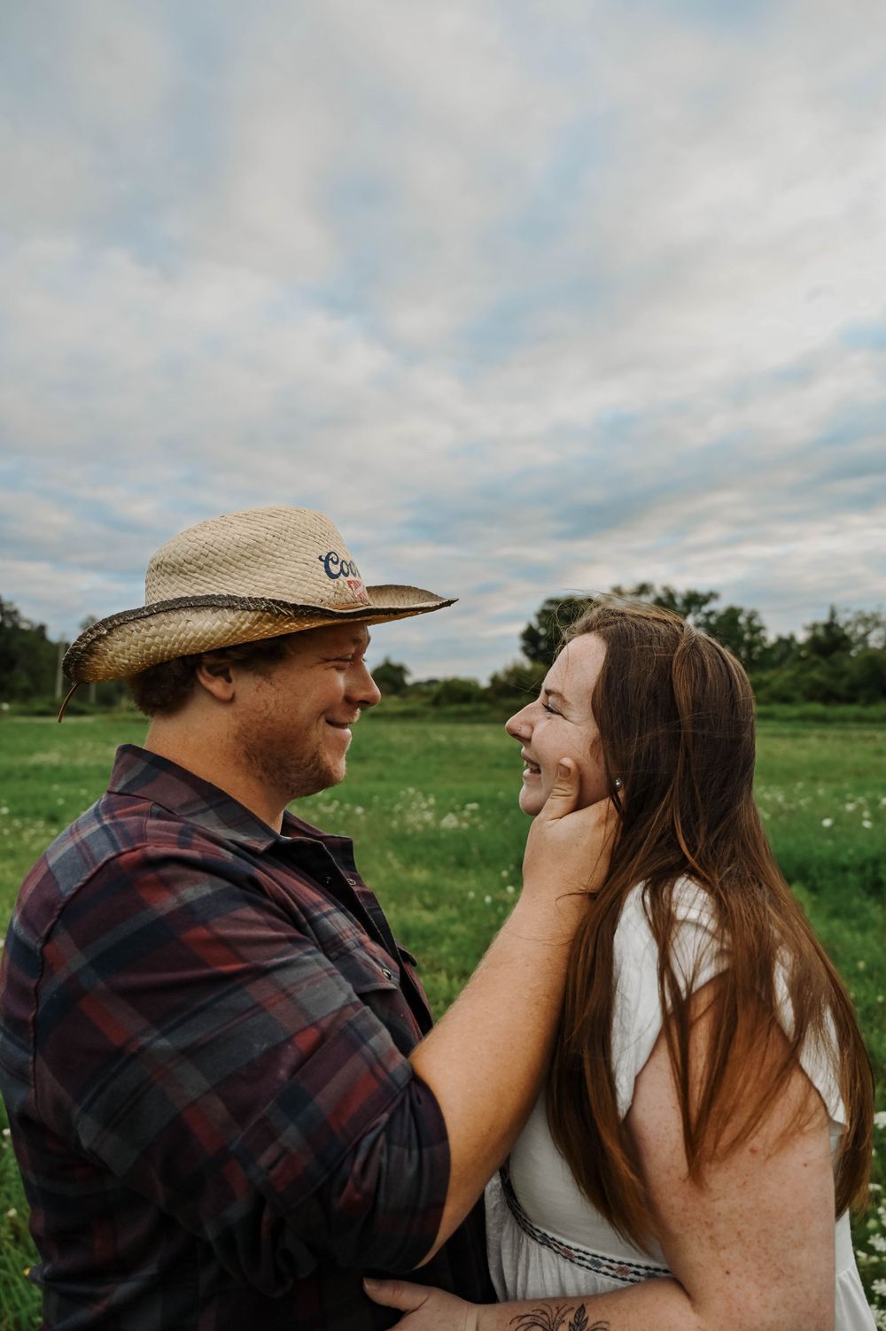 A couple shares an intimate moment together in a field under cloudy skies, with a man wearing a straw hat and plaid shirt.