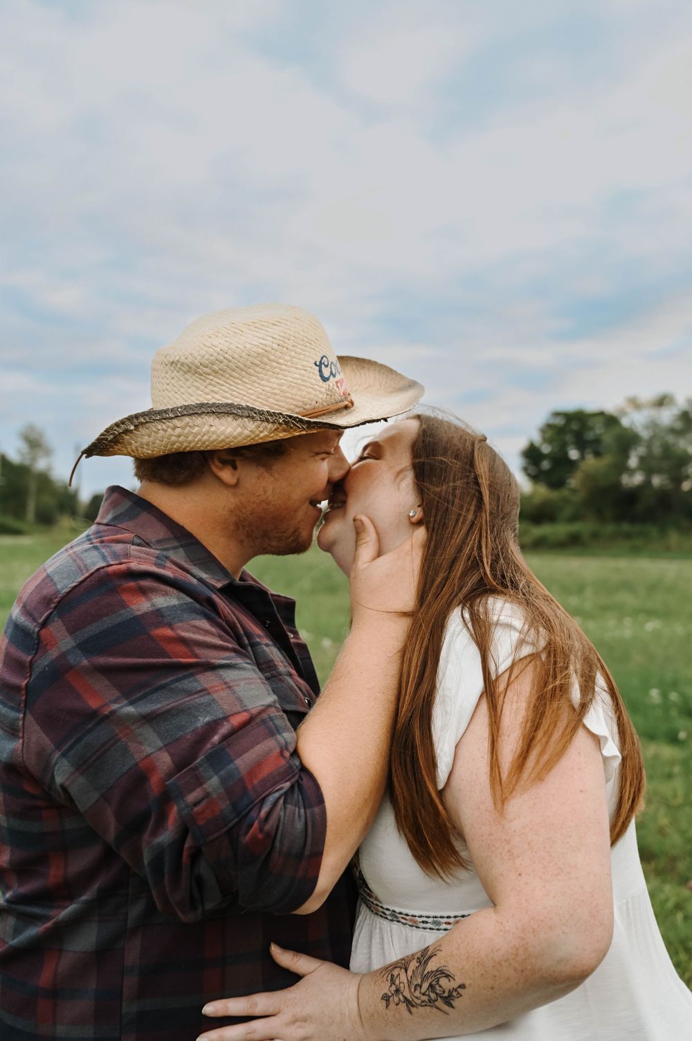 A romantic outdoor photo of a couple embracing in a grassy field, with one wearing a cowboy hat and plaid shirt.
