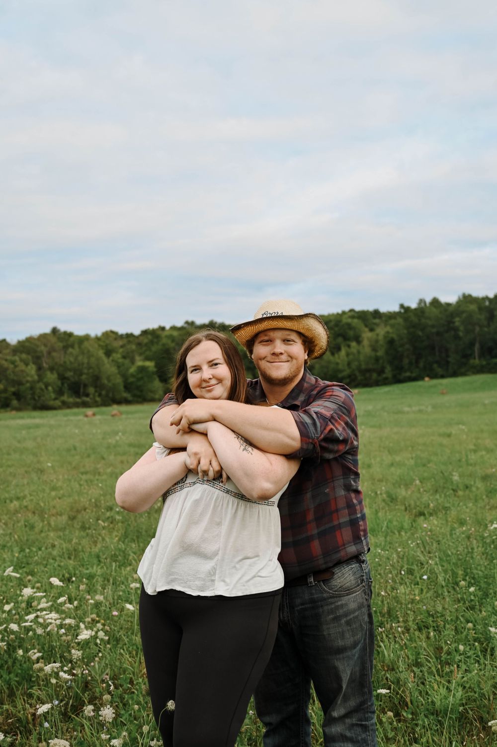 A couple embraces in a sunny meadow with wildflowers and green rolling hills in the background on a summer afternoon.