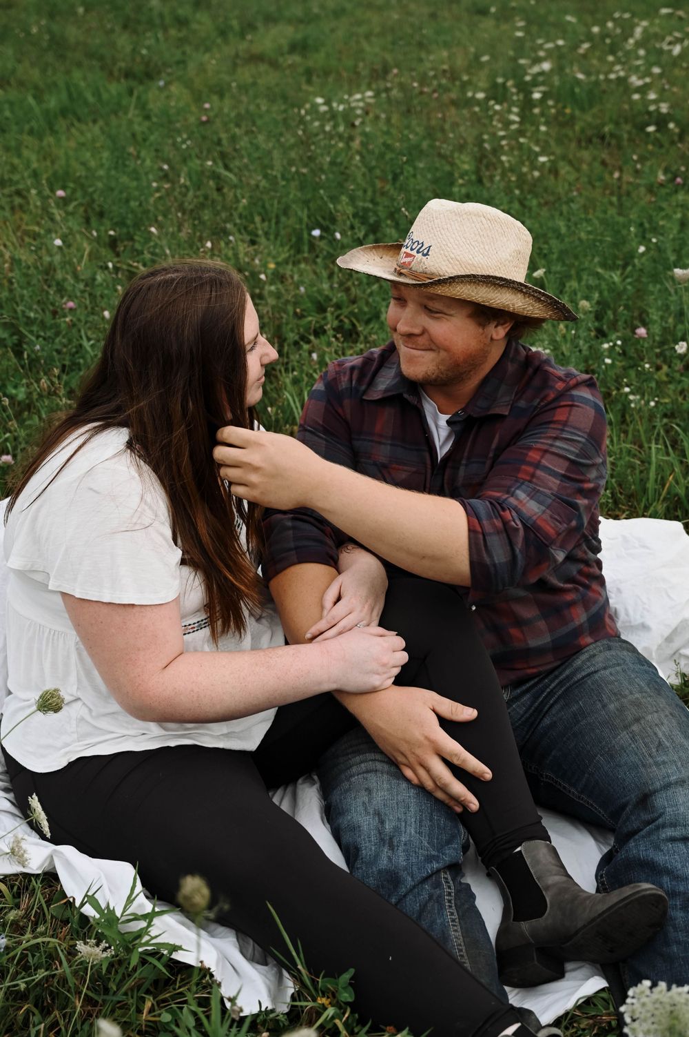 A couple sits together on a white blanket in a grassy meadow with wildflowers on a sunny afternoon.