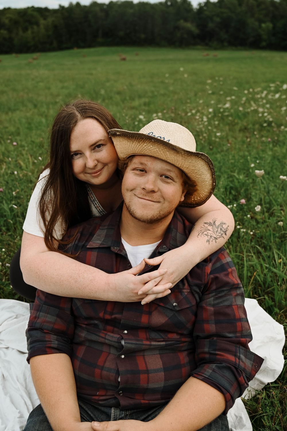 A couple embraces while sitting in a wildflower meadow, with the person in back wearing a plaid shirt and cowboy hat.