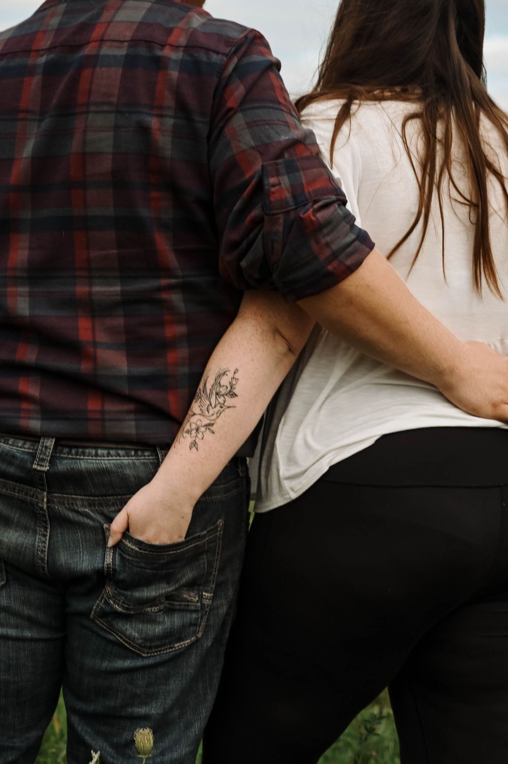 Couple stands close together outdoors showing floral arm tattoo, wearing plaid shirt and white top.