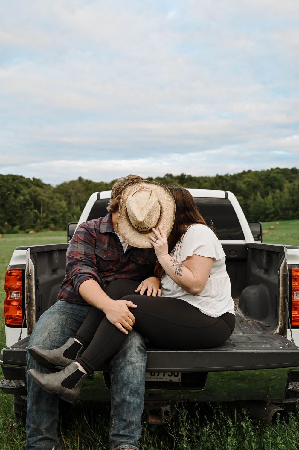 A couple sits close together in the back of a pickup truck parked in a green rural field on a cloudy day.