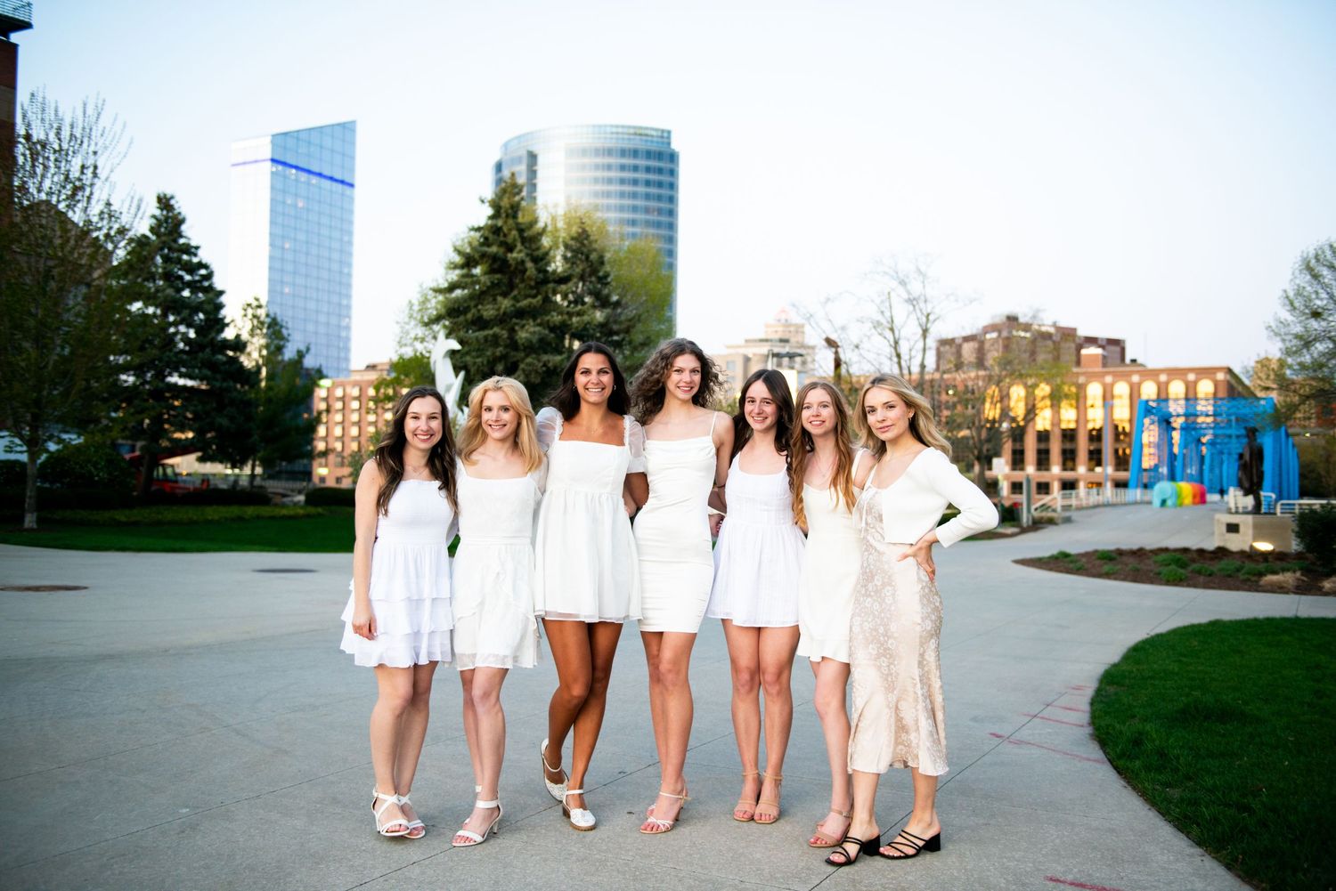 A group in white dresses poses together in a city park with modern skyscrapers and historic buildings in the background.