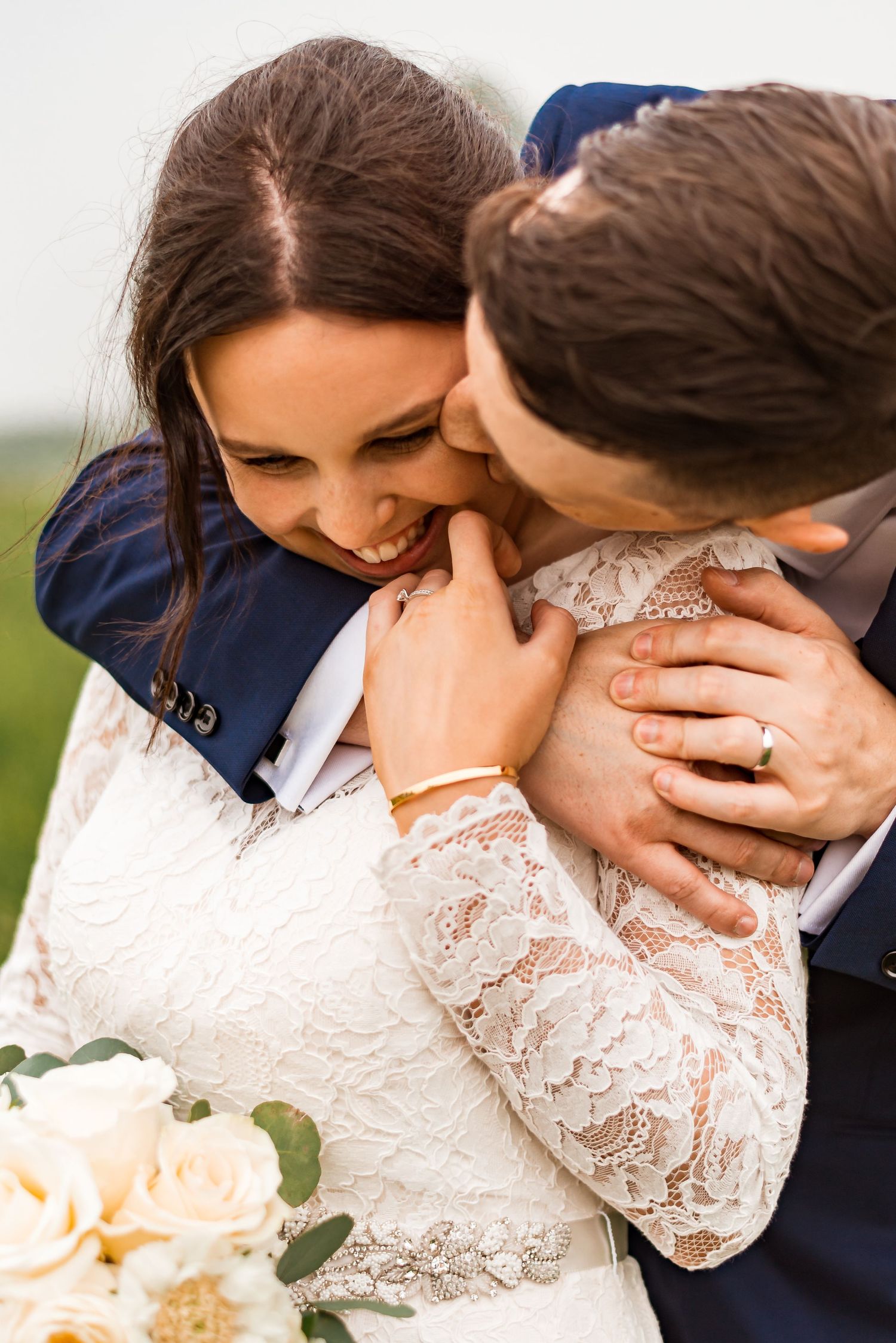 A loving wedding couple embrace outdoors, with the bride wearing a white lace dress and holding cream colored roses.