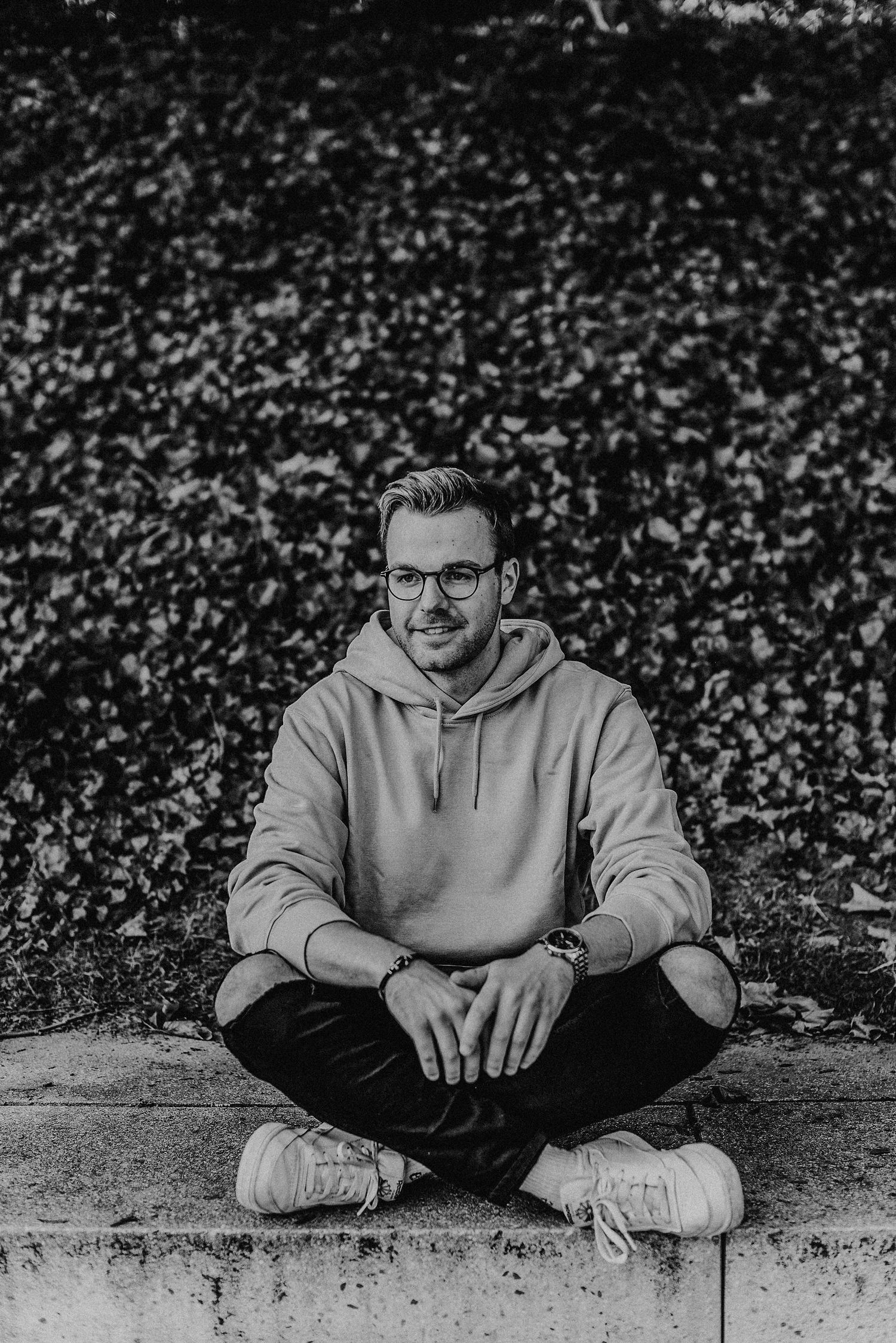 Ein Mann mit Brille und Kapuzenpullover steht lächelnd auf einem Weg, umgeben von Bäumen und Gebäuden im Hintergrund.