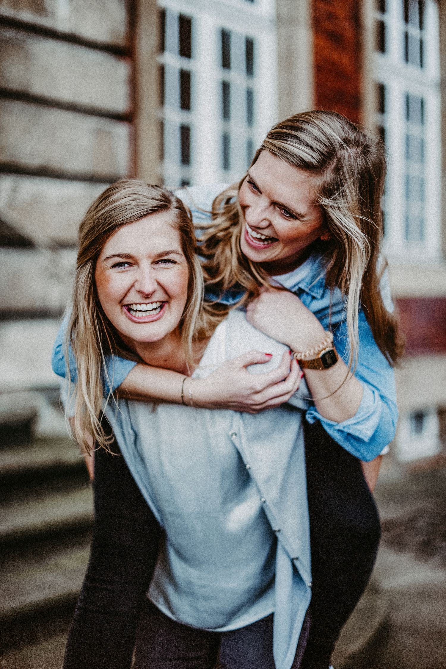 Zwei lachende Frauen, eine trägt die andere auf dem Rücken. Sie stehen vor einem Gebäude mit großen Fenstern.