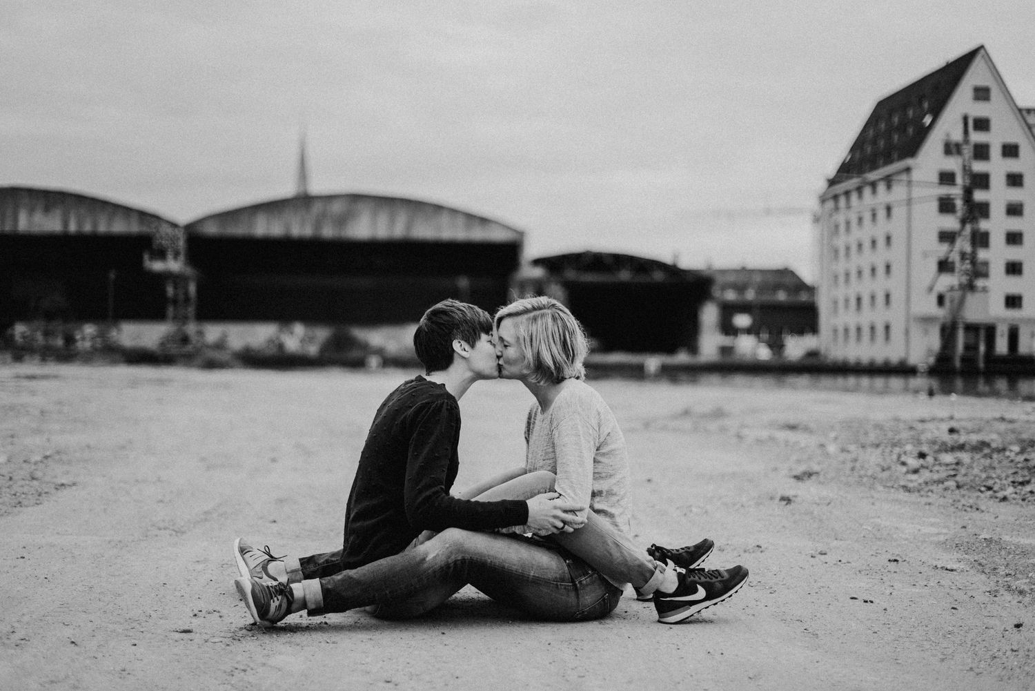 Zwei Personen sitzen auf einem leeren Platz und küssen sich. Im Hintergrund sind große Gebäude und ein bewölkter Himmel.