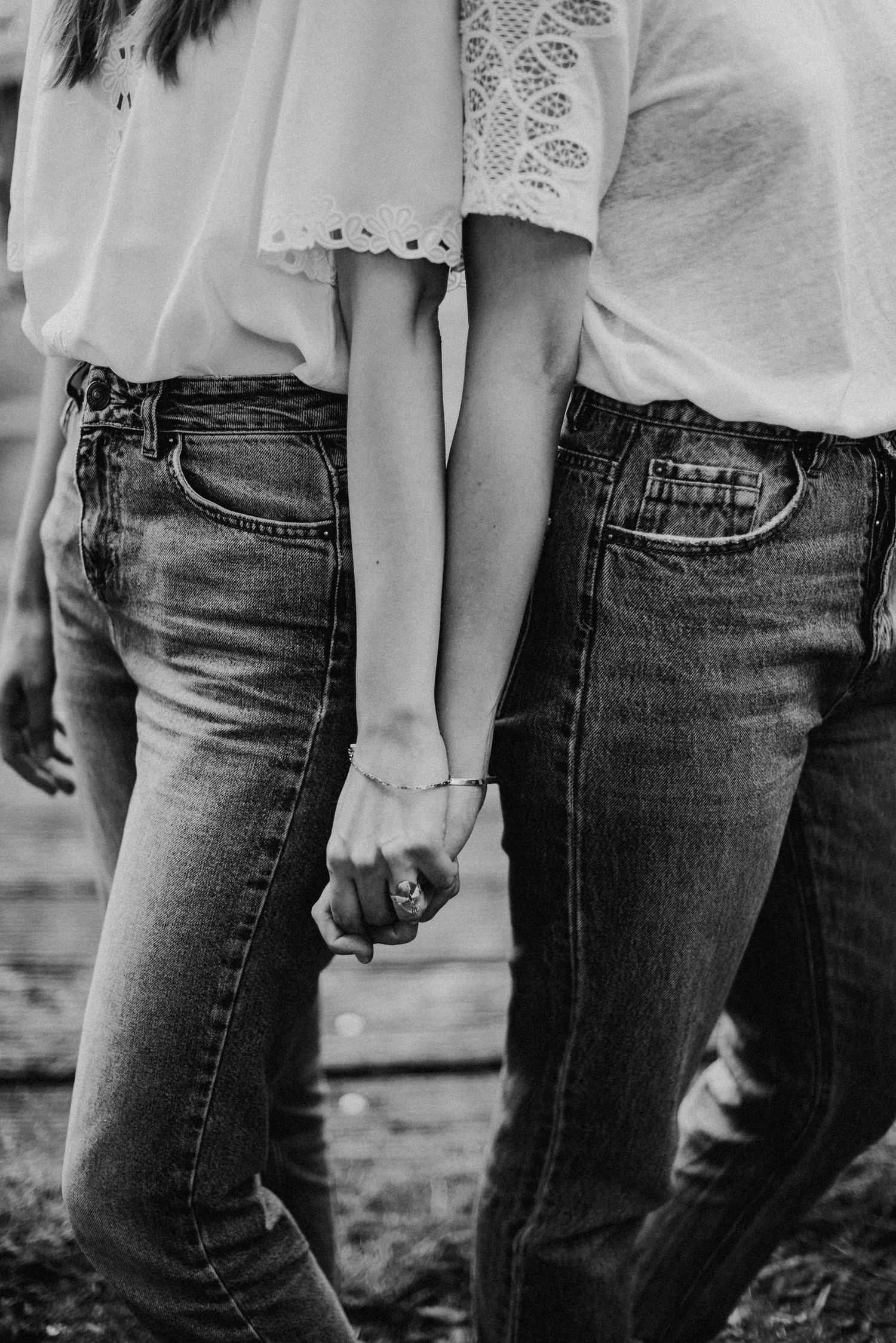 Zwei Personen in Jeans und weißen Oberteilen halten sich an den Händen. Schwarz-weiß-Foto, Fokus auf die Hände und Kleidung.