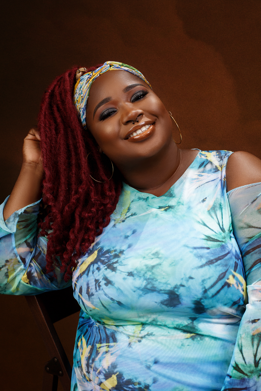Someone in a vibrant blue tie-dye dress with bright red locs and a patterned headscarf poses joyfully against a brown backdrop.