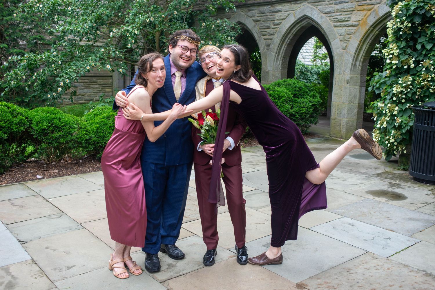 A playful group in formal attire poses together in front of a stone archway in a garden setting.