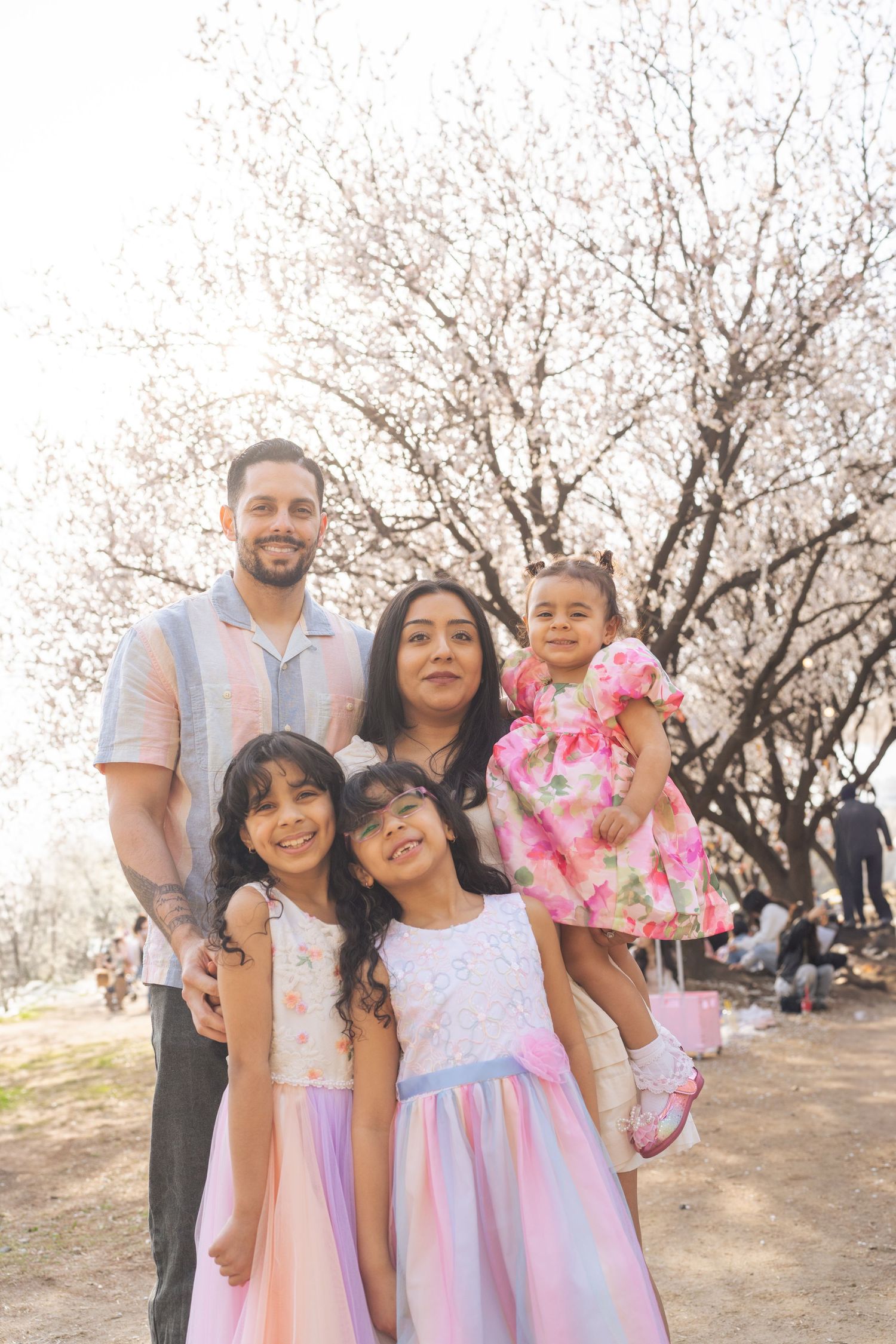 Family wearing coordinated pink and purple outfits poses together among blooming cherry blossom trees in a park.