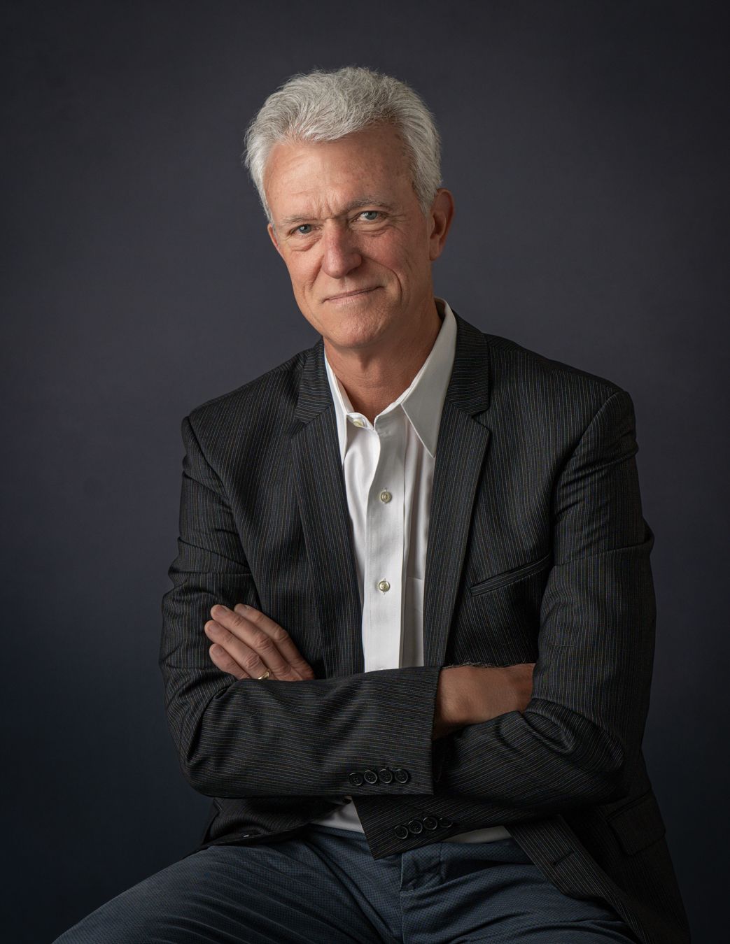 Professional man with gray hair in dark blazer sits with arms crossed against dark background. Best photographer Williamsburg VA Headshot