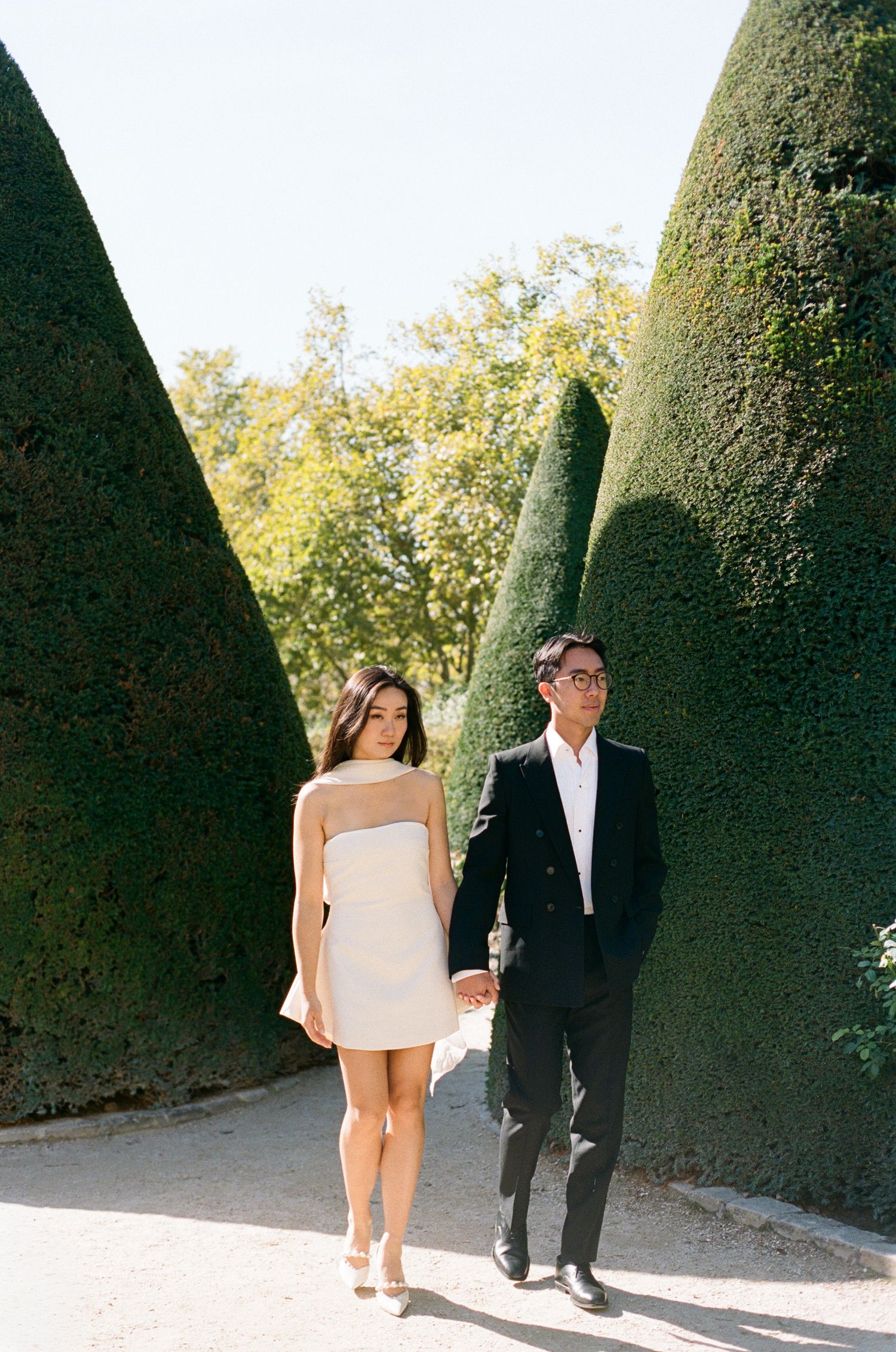 A couple in formal attire walks along a path bordered by tall manicured hedges in a garden setting.