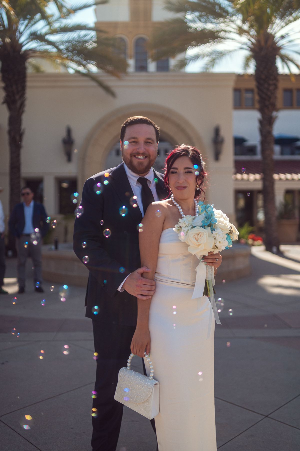 A bride in a white dress and groom in a dark suit pose together with bubbles floating around them at a venue with palm trees.