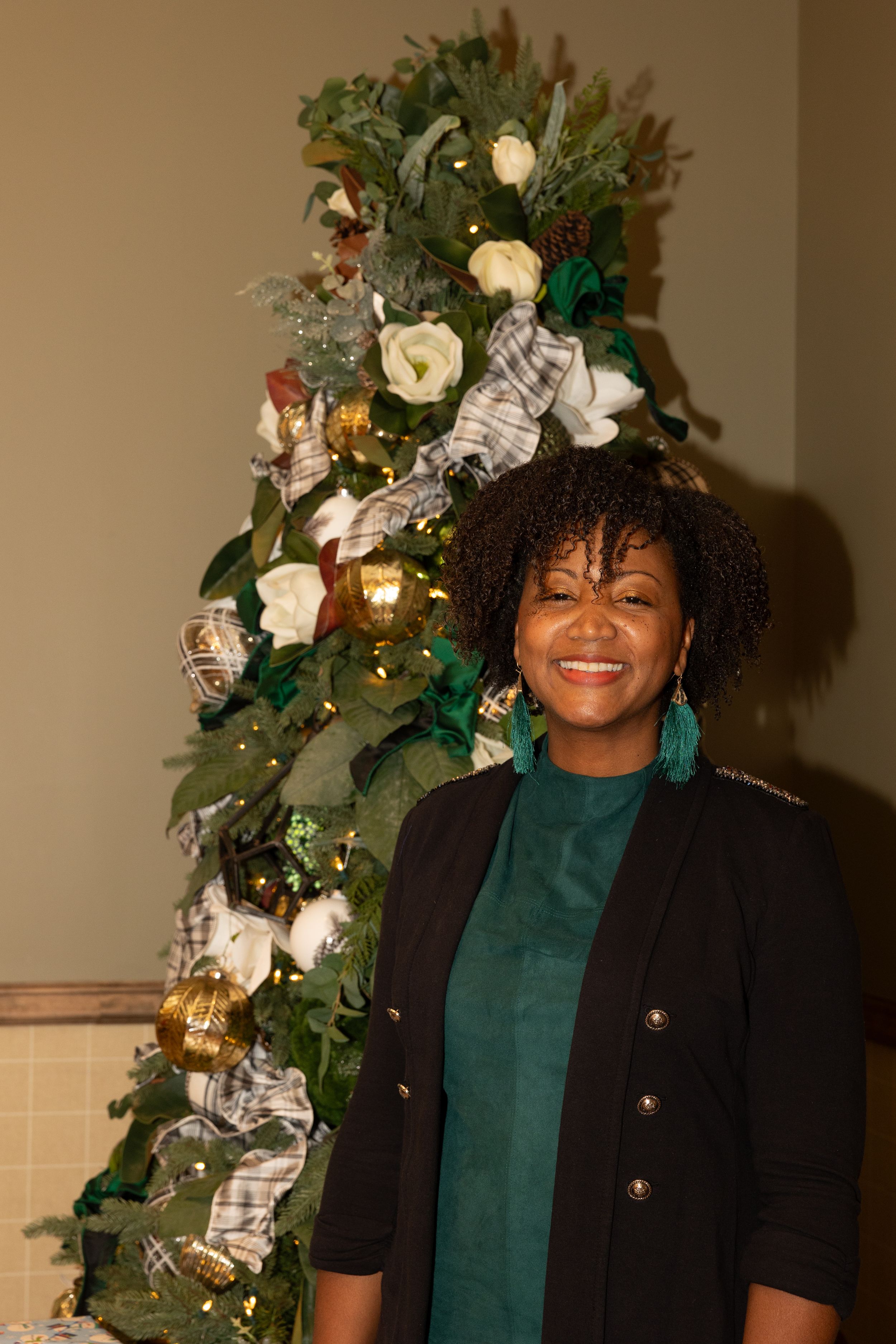 Person wearing black and green attire poses in front of decorated Christmas tree with white flowers and ornaments.