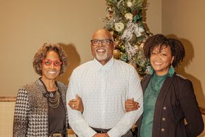 A group of people stand together smiling at a holiday gathering with a decorated Christmas tree in the background.