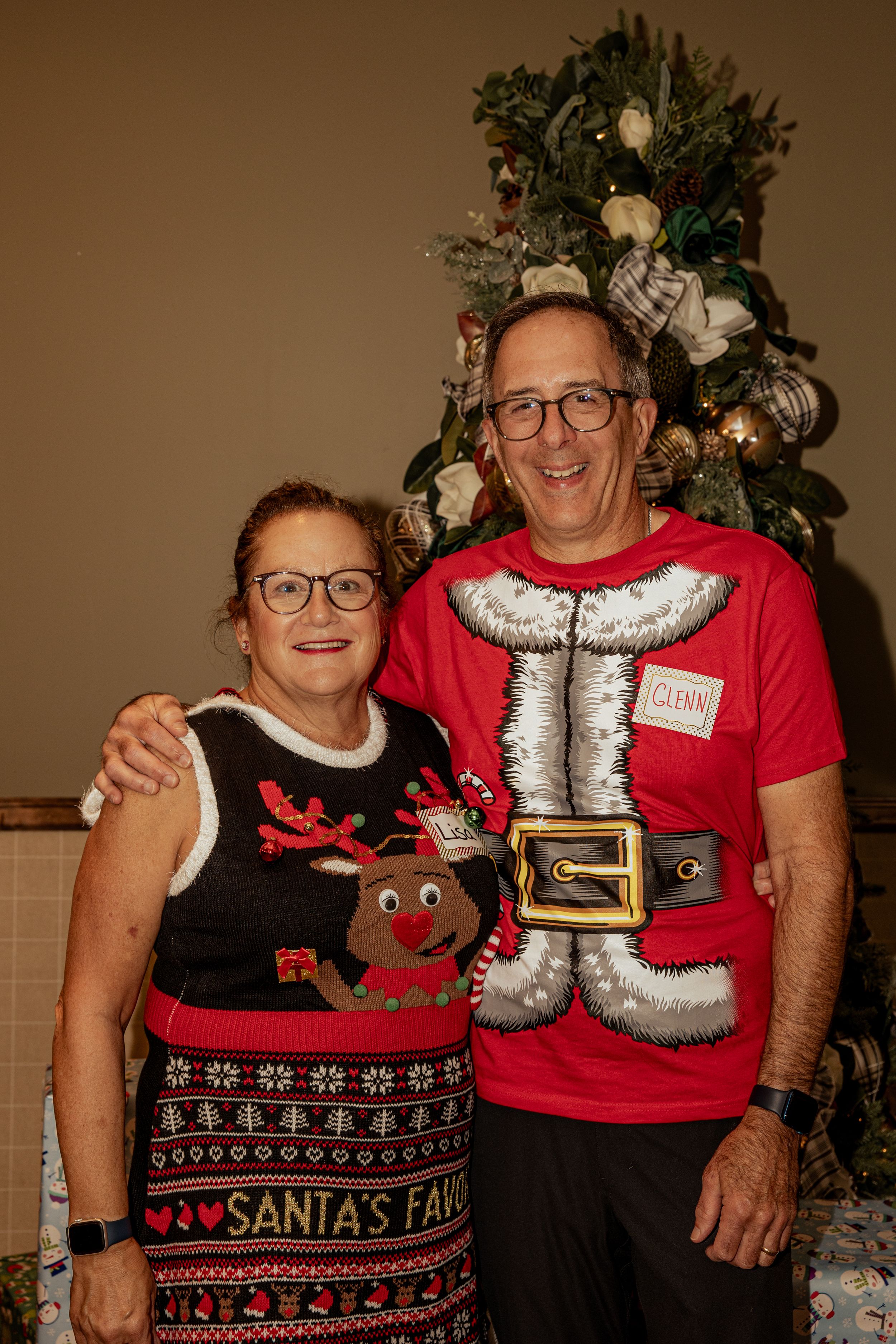 Two people wearing festive Christmas sweaters with reindeer designs pose together near a decorated holiday tree.