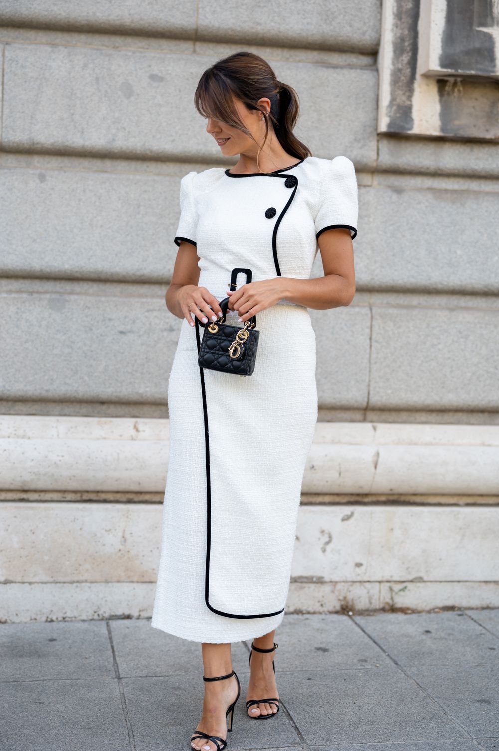 Woman in white dress with black trim and sandals walking on a city sidewalk near stone building steps.
