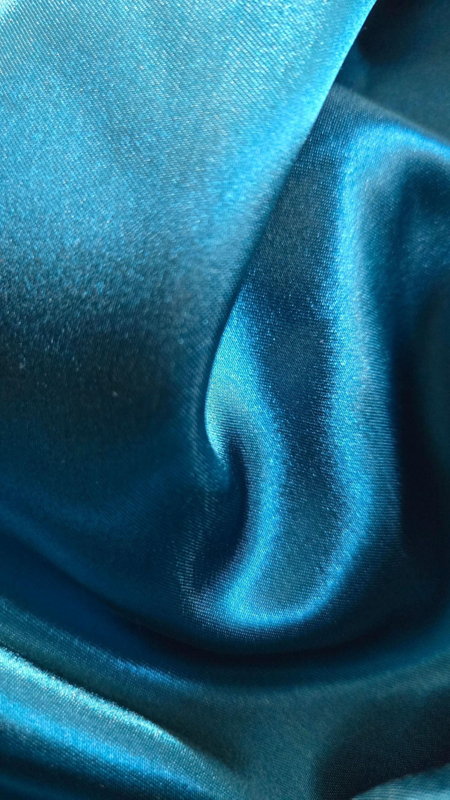 Flowing waves of shiny teal silk fabric create an elegant rippled texture in deep turquoise blue tones.