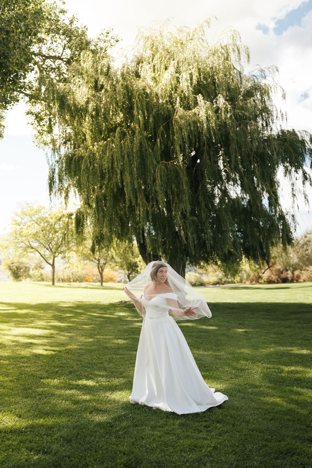 A bride in a flowing white dress poses gracefully on a lawn with weeping willow trees.