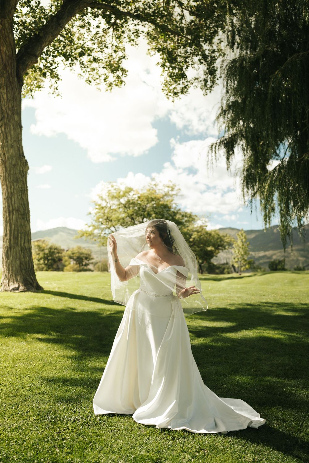 Bride spins gracefully in her white wedding gown on a sunny lawn with willow trees.