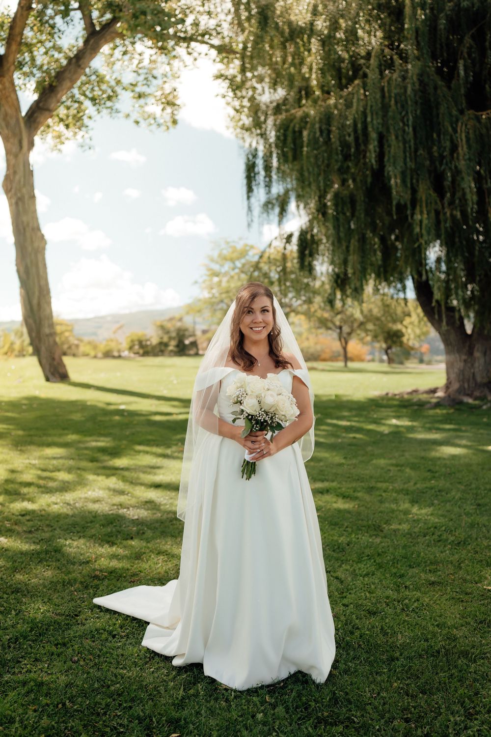 White wedding dress flows across green lawn under willow trees on sunny day.