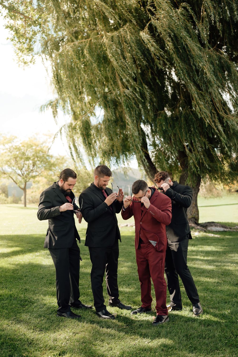 Groomsmen in formal attire perform synchronized dance moves under tree branches.
