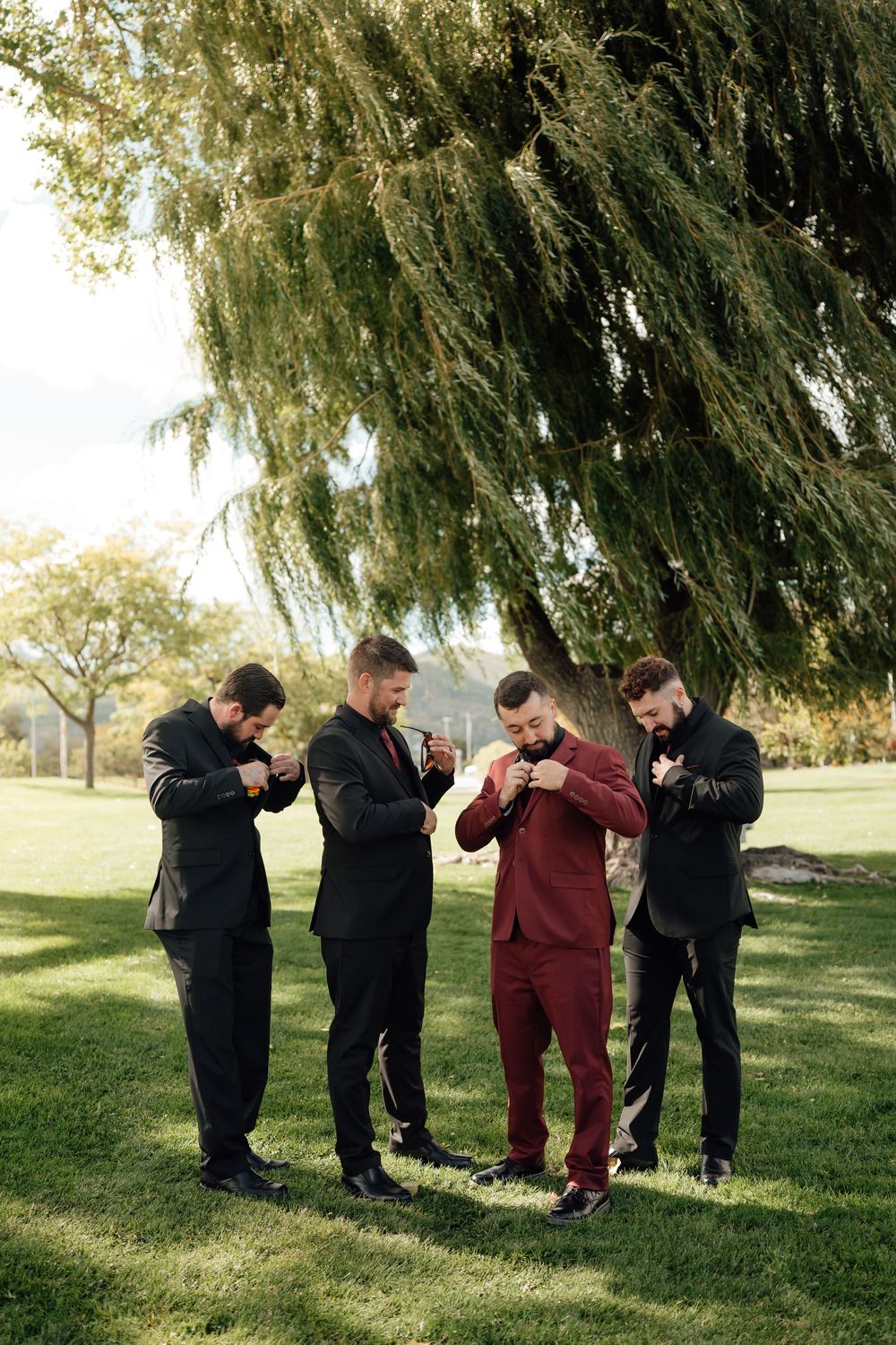 Groomsmen in burgundy and black suits pose playfully under a willow tree.
